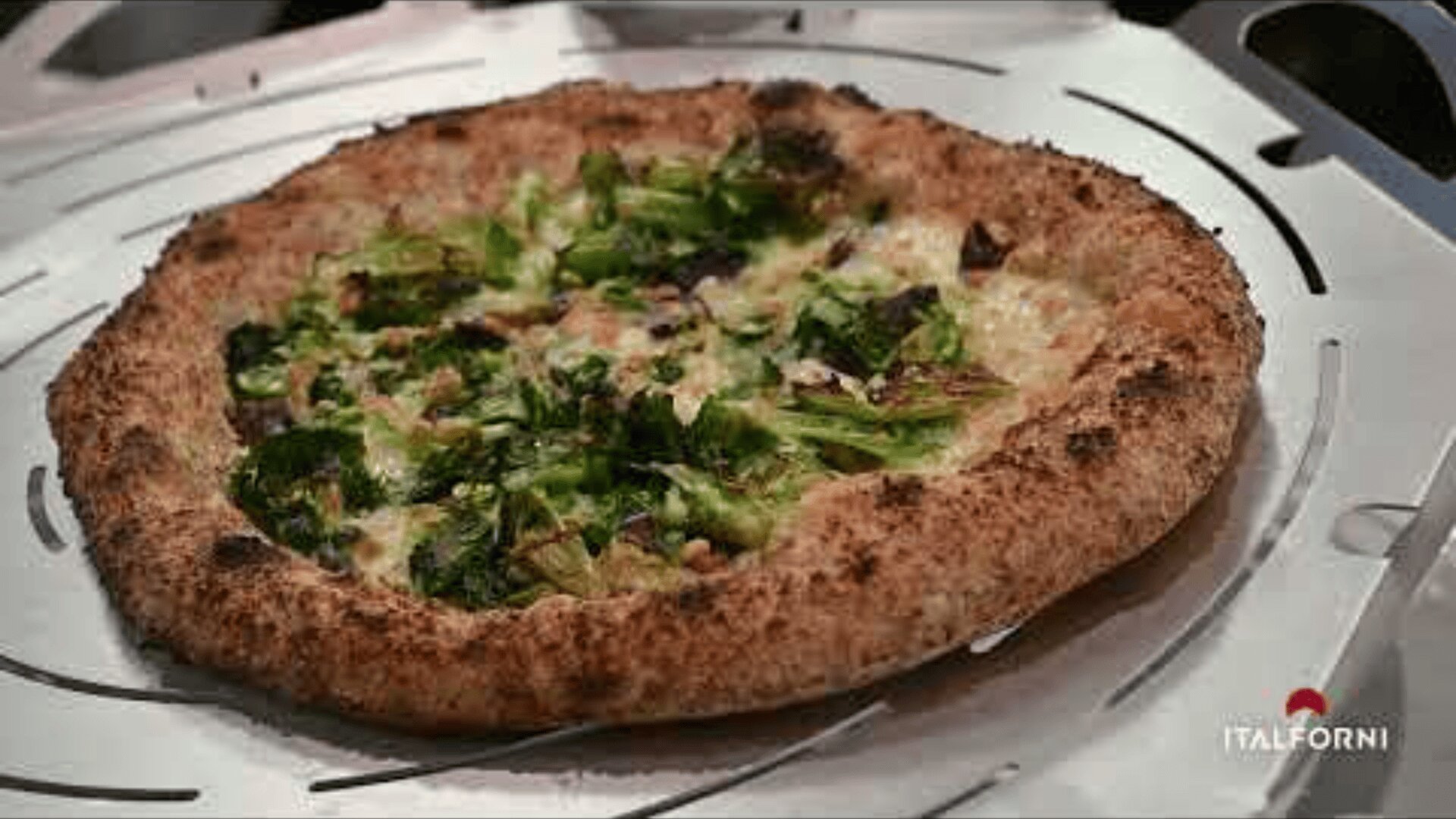 A wood-fired pizza topped with greens and cheese sits on a metal tray. The pizza has a thick, golden-brown crust and is displayed on an Italforni oven surface.