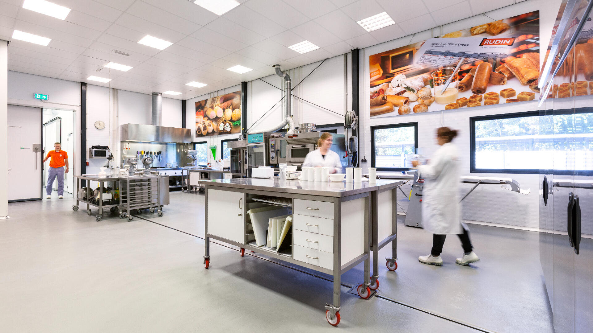 A bright, modern commercial kitchen with stainless steel equipment, large posters of baked goods on the walls, and people in white lab coats working and walking around.