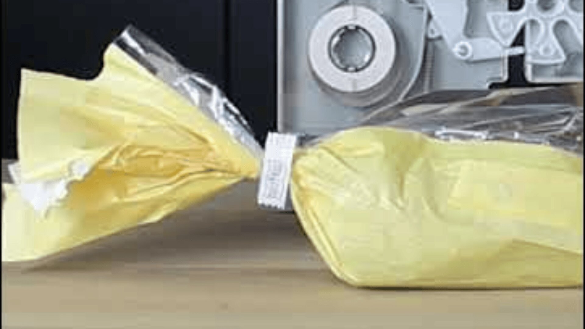 A yellow plastic bag, sealed with a white ribbon, lies on a wooden base, with a gray mechanical device in the background.