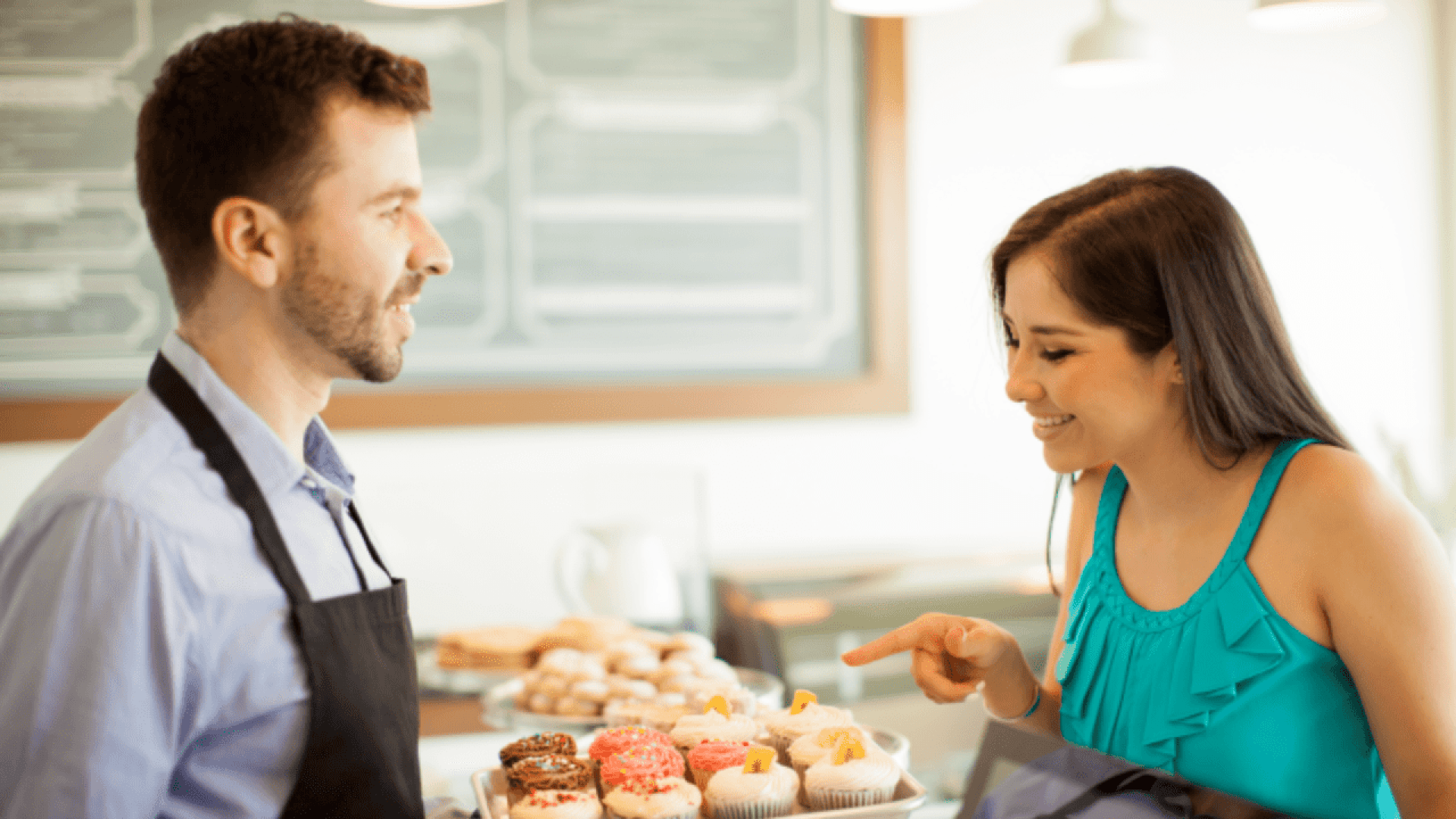 Eine lächelnde Frau in einem türkisfarbenen Oberteil zeigt auf bunte Muffins auf einem Tablett, das ein Mann in einer schwarzen Schürze hinter einer Bäckertheke hält. Sie scheinen ein freundliches Gespräch zu führen.
