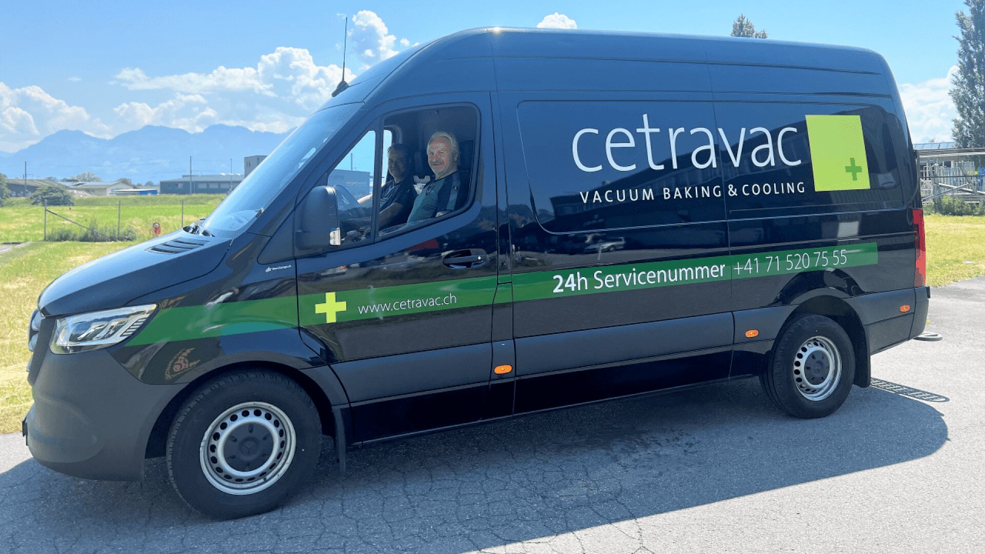 A dark van with Cetravac Vacuum Baking & Cooling branding is parked on a sunny road. Two people inside smile and give thumbs up. The mountains and a grassy field are visible in the background.
