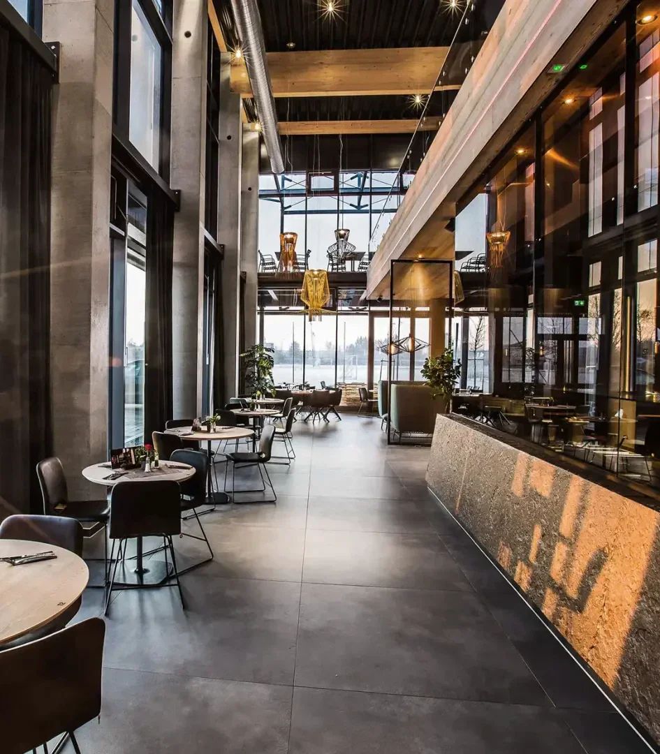 Modernes Restaurantinterieur mit hohen Decken, großen Fenstern, stilvoller Beleuchtung und modernen Möbeln. Auf der linken Seite befinden sich Tische und Stühle, auf der rechten Seite ein langer Bartresen, in den natürliches Licht fällt.