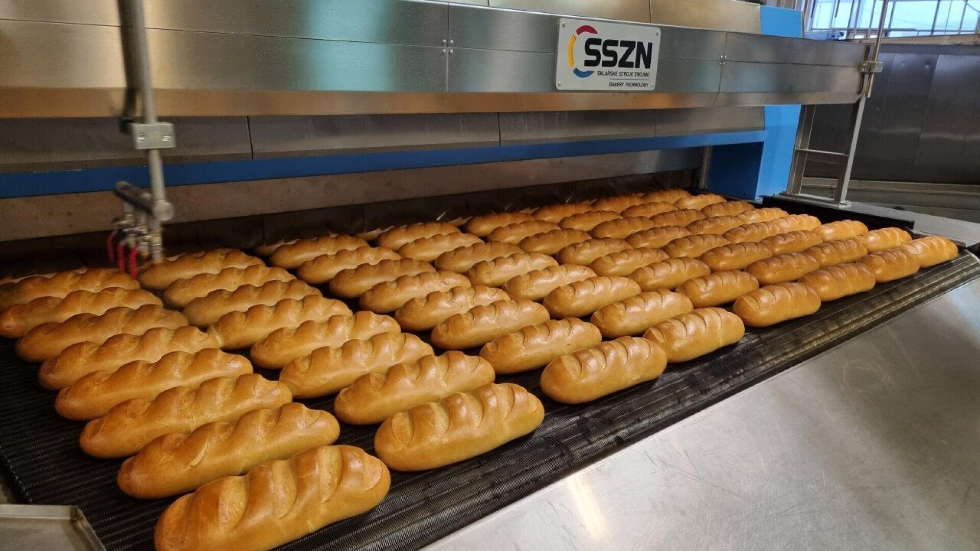 Reihen von frisch gebackenen, geflochtenen Brotlaiben laufen über ein Förderband in einer industriellen Bäckerei. Über den Broten steht eine große Edelstahlmaschine mit der Aufschrift SSZN.