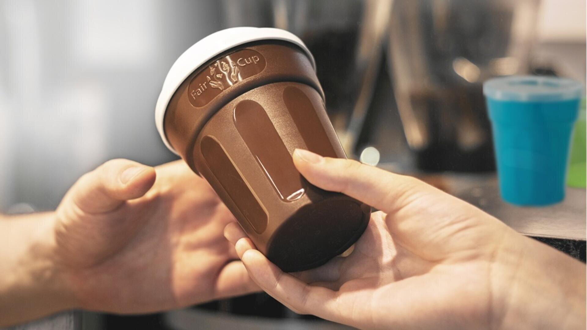 A hand passes a brown reusable coffee cup with a white lid and embossed logo to another hand, with blurred kitchen items in the background.