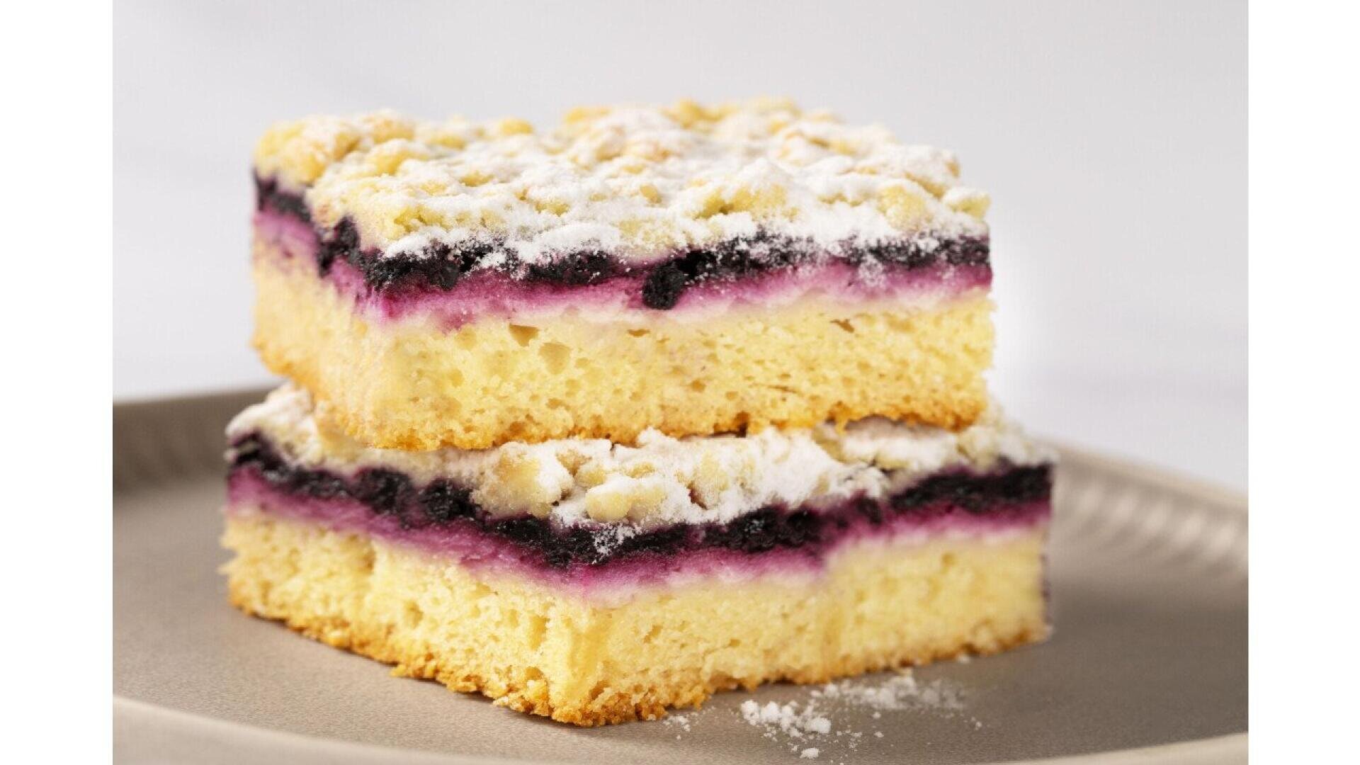 Two square pieces of cake with a yellow base, a purple berry filling, and a crumbly topping dusted with powdered sugar, stacked on a beige plate.