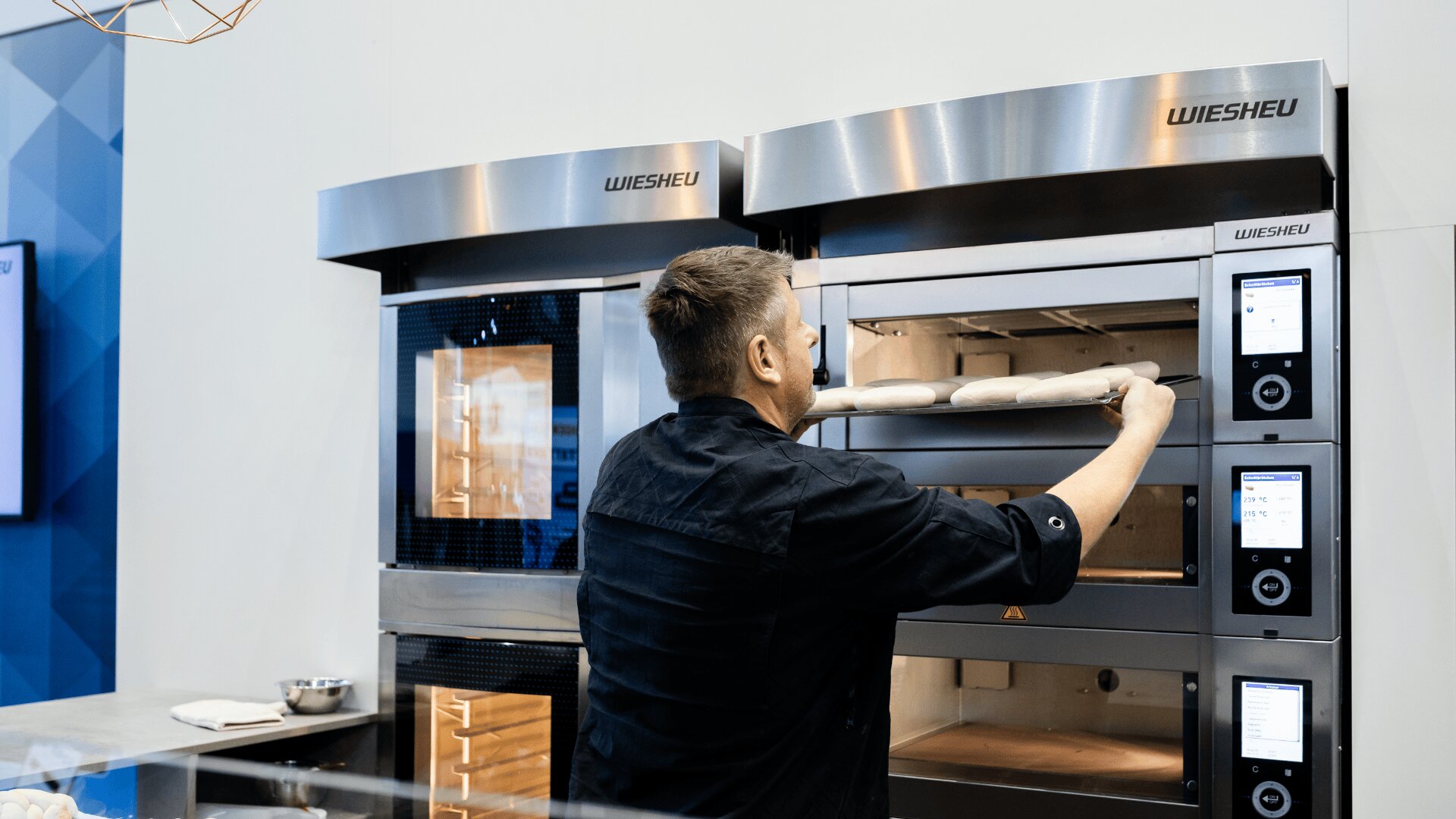 Ein Mann in einer schwarzen Kochjacke schiebt ein Tablett mit ungebackenen Baguettes in einen Großofen in einer modernen Bäckereiküche.