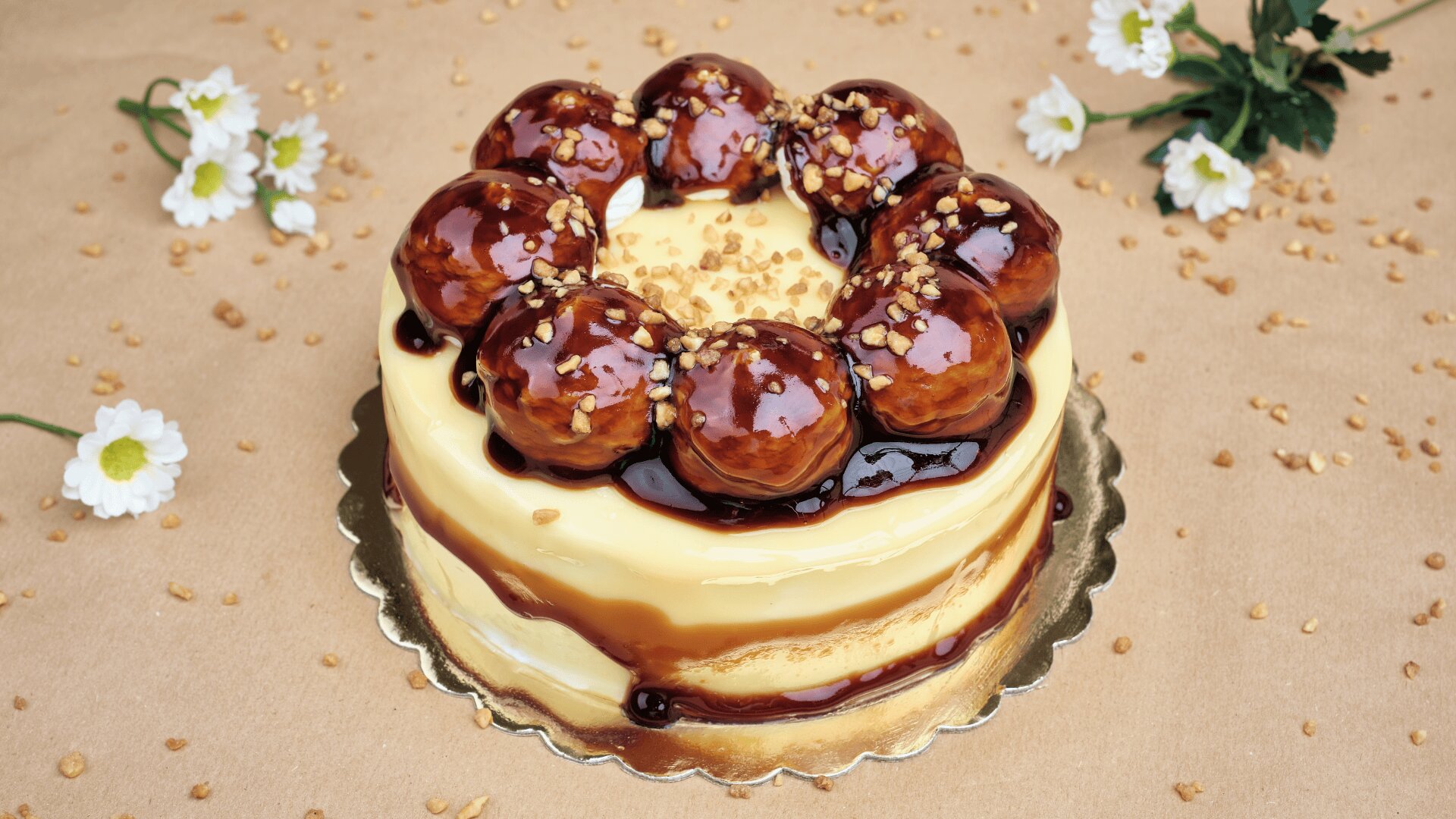 A round layered cake topped with caramel-glazed cream puffs, chocolate sauce, and chopped nuts. The cake sits on a gold base with white flowers and nut pieces scattered around on a tan surface.