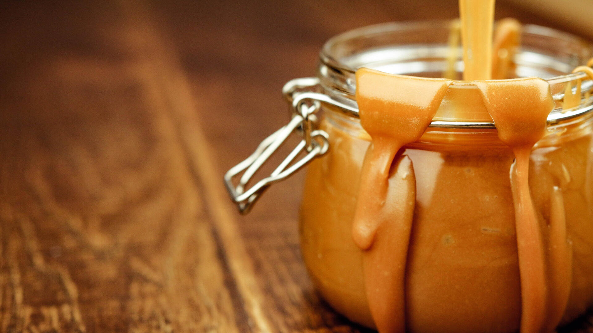 Auf einer Holzoberfläche steht ein Glasgefäß mit dicker Karamellsauce. An den Seiten tropft Karamell herunter, und von oben wird mehr hineingegossen.