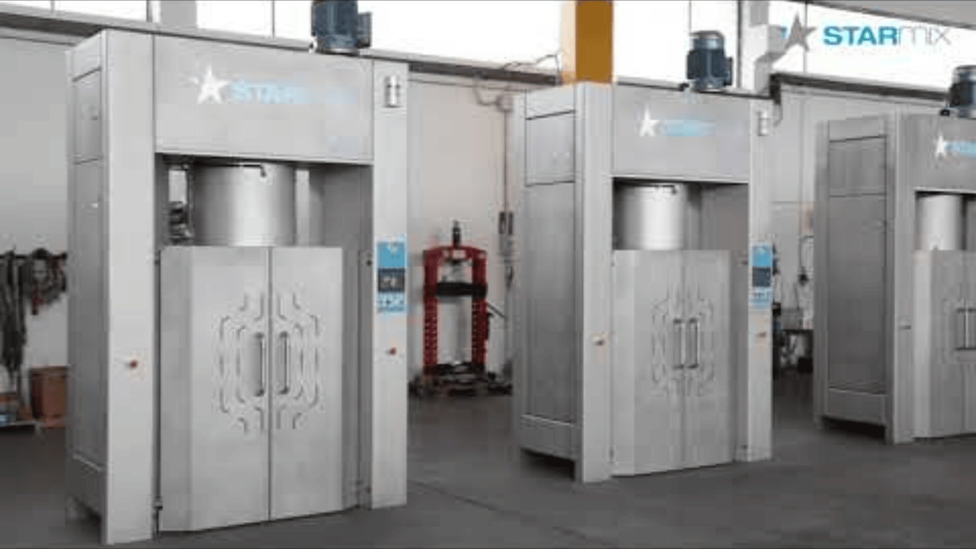 Drei große Industriemaschinen aus Metall mit geschlossenen Türen stehen nebeneinander in einer Fabrikhalle, in der im Hintergrund verschiedene Geräte und Werkzeuge zu sehen sind.