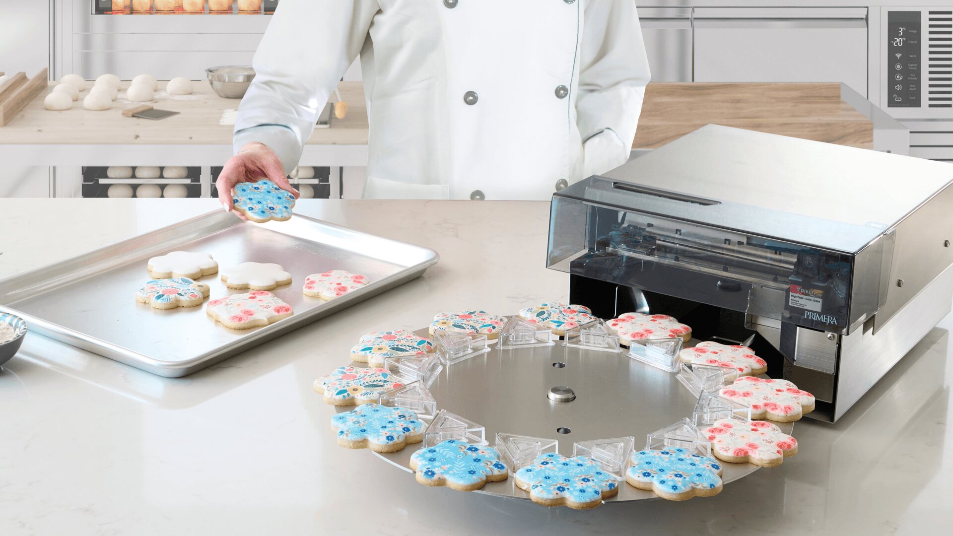 A baker in a white chef’s coat holds a decorated cookie over a tray, while a machine prints blue and pink designs onto flower-shaped cookies arranged on a rotating platter.