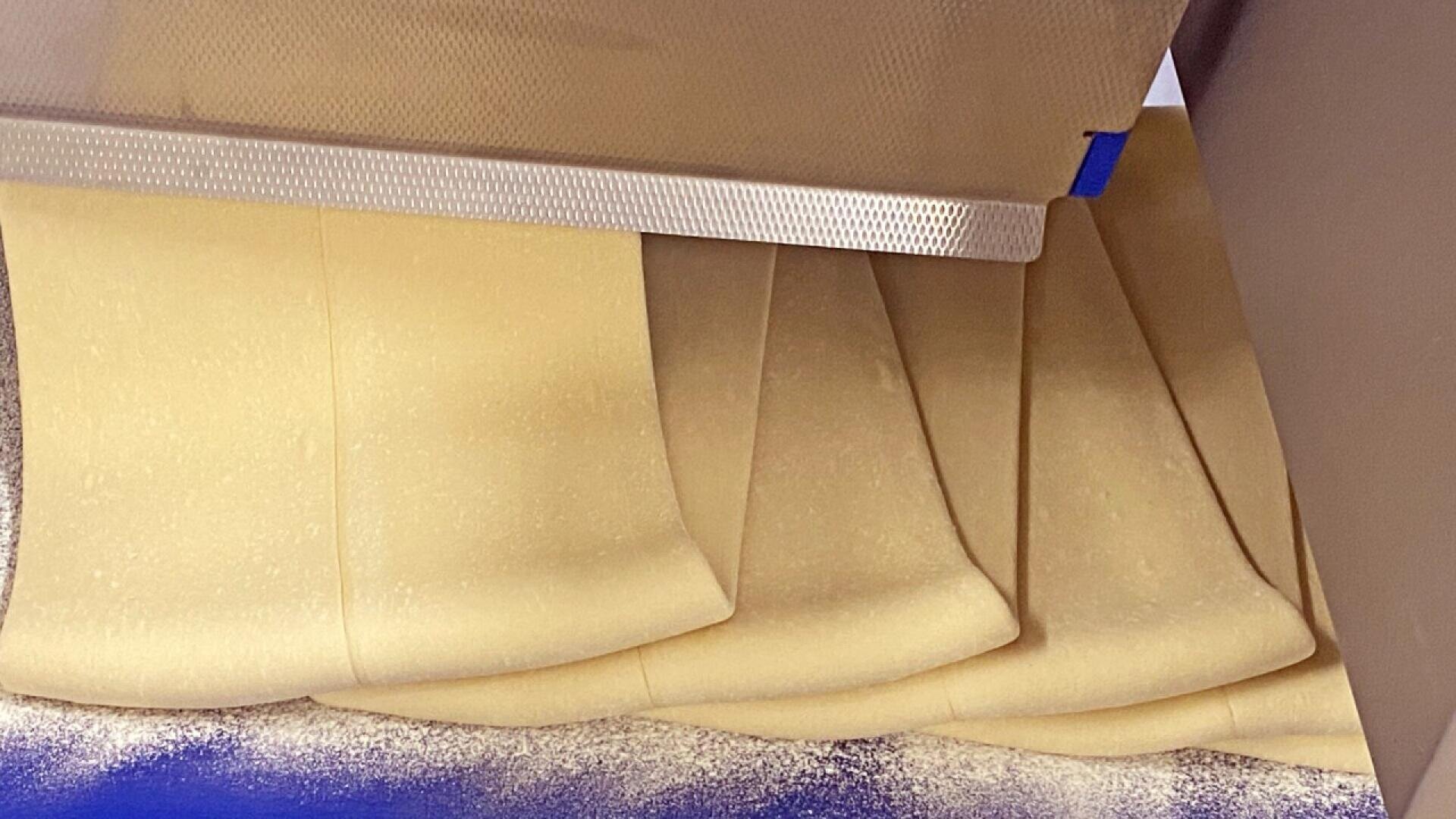 Die Teigblätter werden von einer Maschine geschichtet und gefaltet, wobei auf der Oberfläche ein Mehlstaub zu sehen ist.
