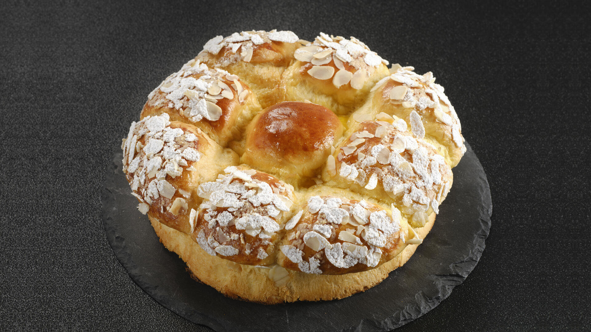 Ein rundes, goldfarbenes süßes Brot, belegt mit Mandelscheiben und Puderzucker, auf einer dunklen Schieferplatte vor schwarzem Hintergrund.