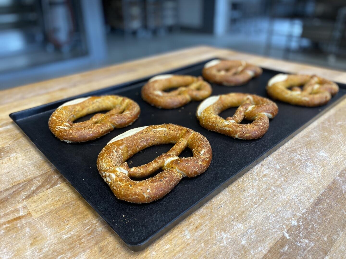 Ein schwarzes Backblech mit sechs frisch gebackenen Laugenbrezeln steht auf einer Holztheke in einer Bäckereiküche. Die Brezeln sind goldbraun und mit grobem Salz bestreut.