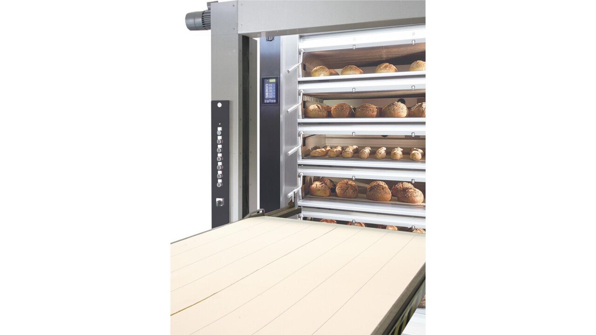 A commercial bakery oven with multiple racks filled with trays of round loaves of bread, next to a conveyor belt for moving baked goods.