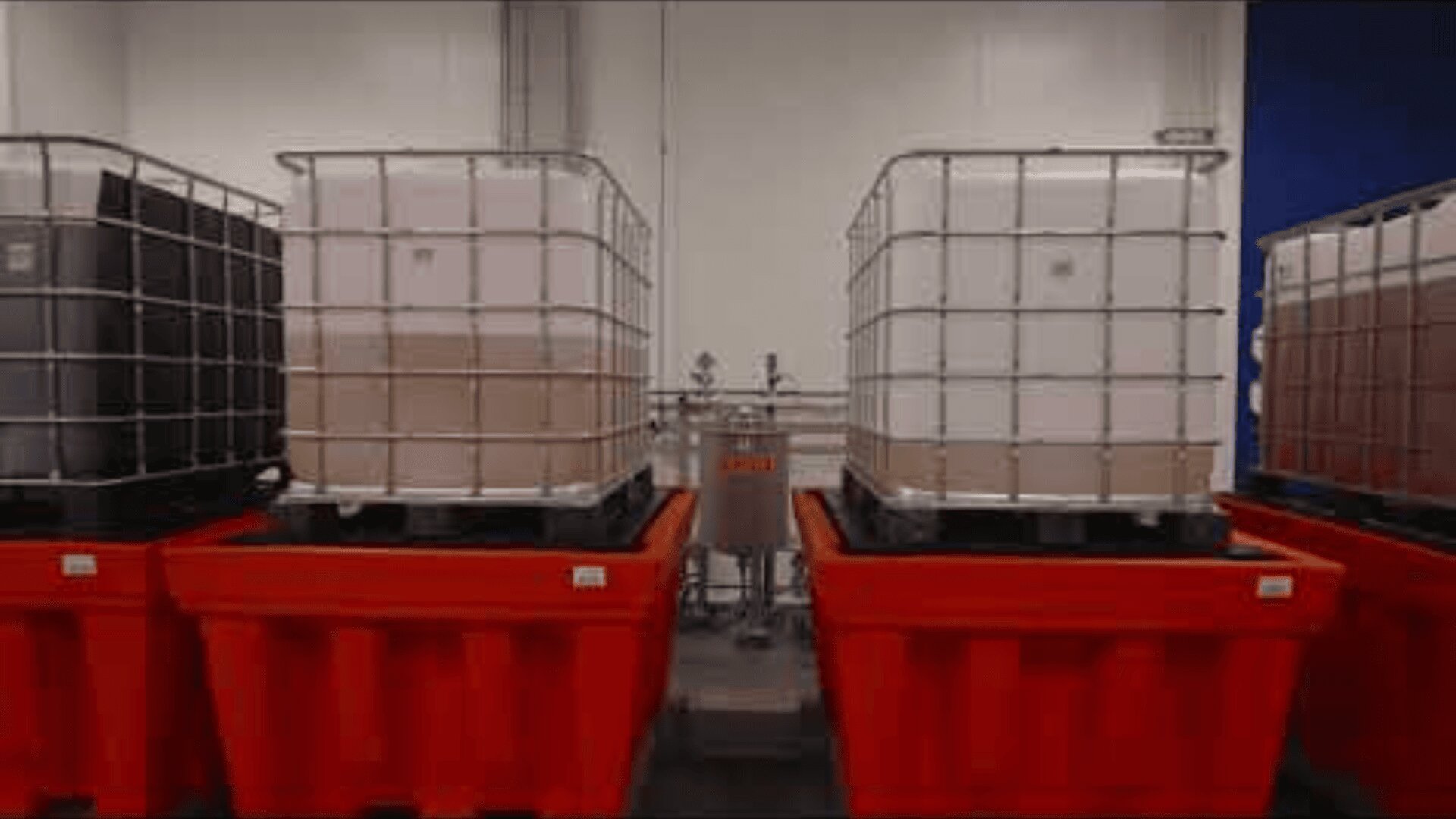 Two large white industrial fluid containers stand on bright red collection pallets in a clean hall with pipes and equipment visible in the background.