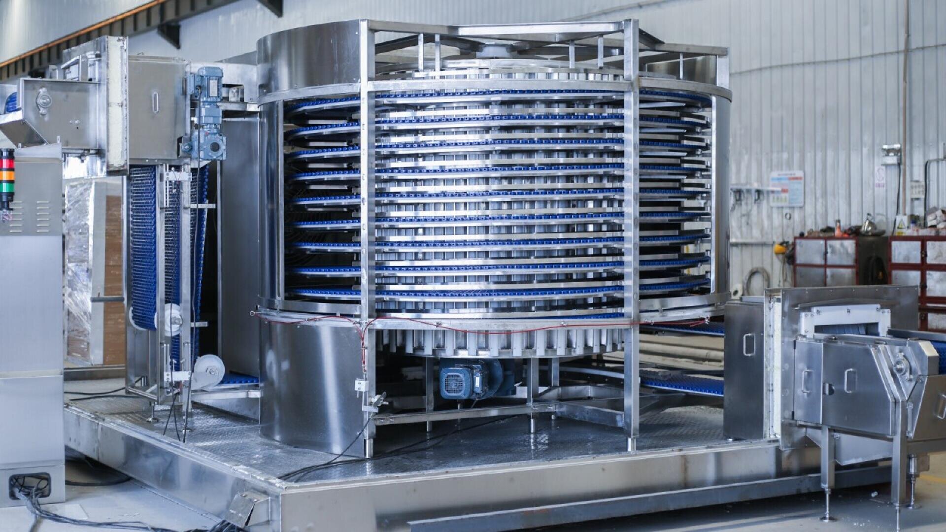 Ein großer industrieller Spiralgefrierschrank aus Edelstahl mit mehreren blauen Förderbändern steht in einer Fabrik mit Metallwänden und verschiedenen Bedienfeldern.