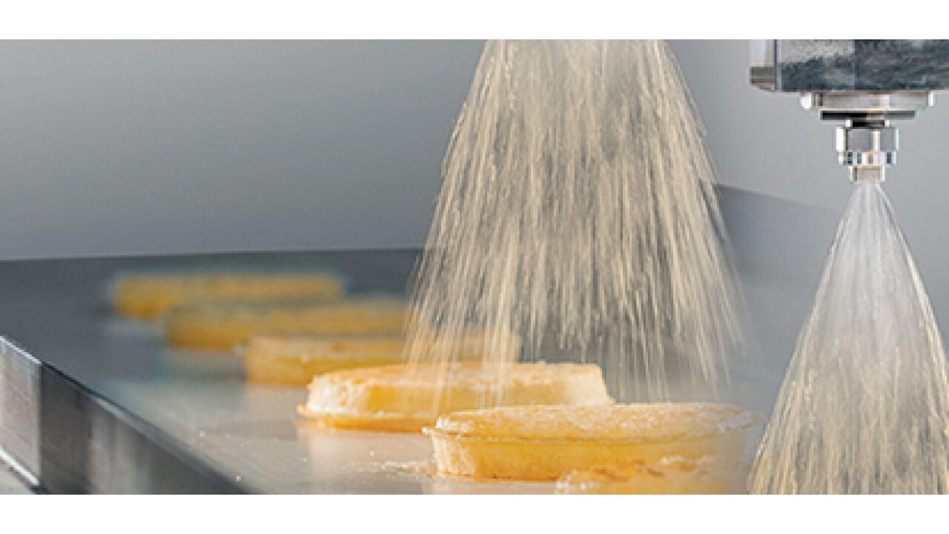 A close-up of food products, possibly pastries, moving on a conveyor belt while being sprayed with a fine mist or coating from a metal nozzle in a food processing facility.