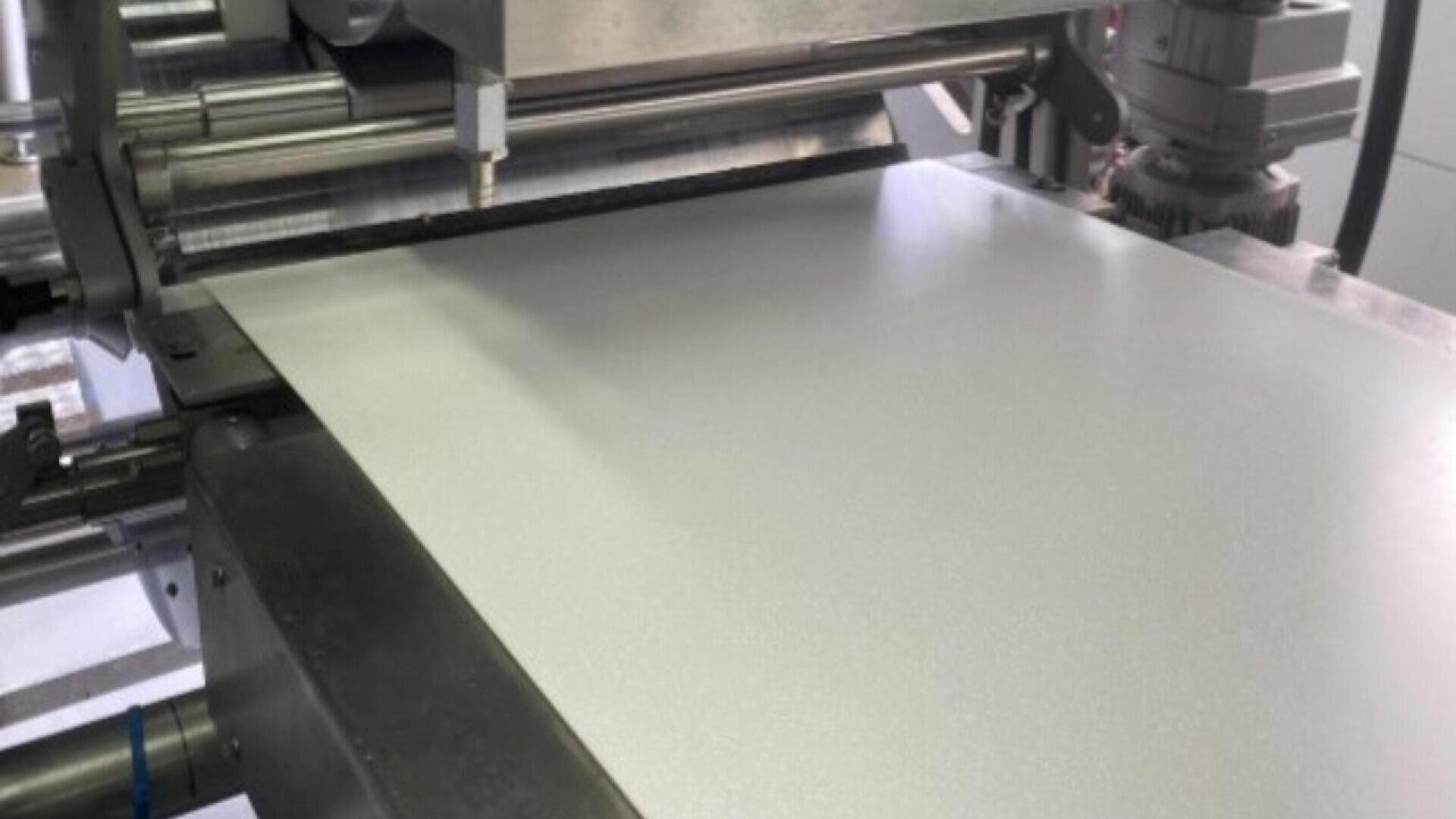 Eine weiße Platte wird in einer Fabrik von einer Industriemaschine mit Metallrollen und -teilen bearbeitet.