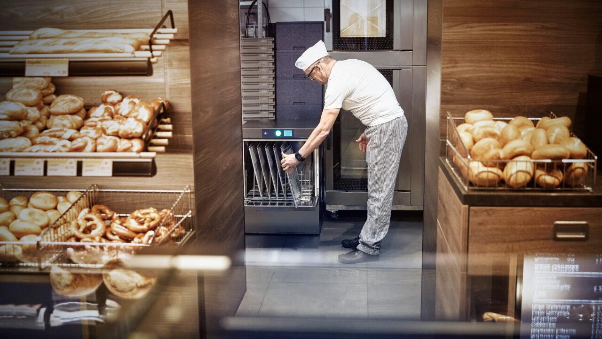 An elderly baker wearing a white shirt, cap, and checkered pants operates a machine in a bakery, surrounded by shelves filled with various breads and pastries.