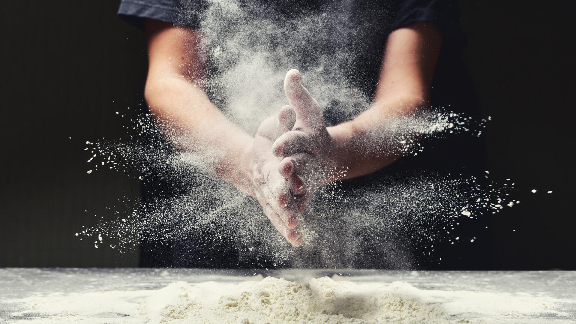 Eine Person klatscht über einer mit Mehl bestäubten Fläche in die mehlbedeckten Hände und erzeugt so eine dramatische Pulverwolke in der Luft. Der dunkle Hintergrund hebt die in der Luft schwebenden Mehlpartikel hervor.