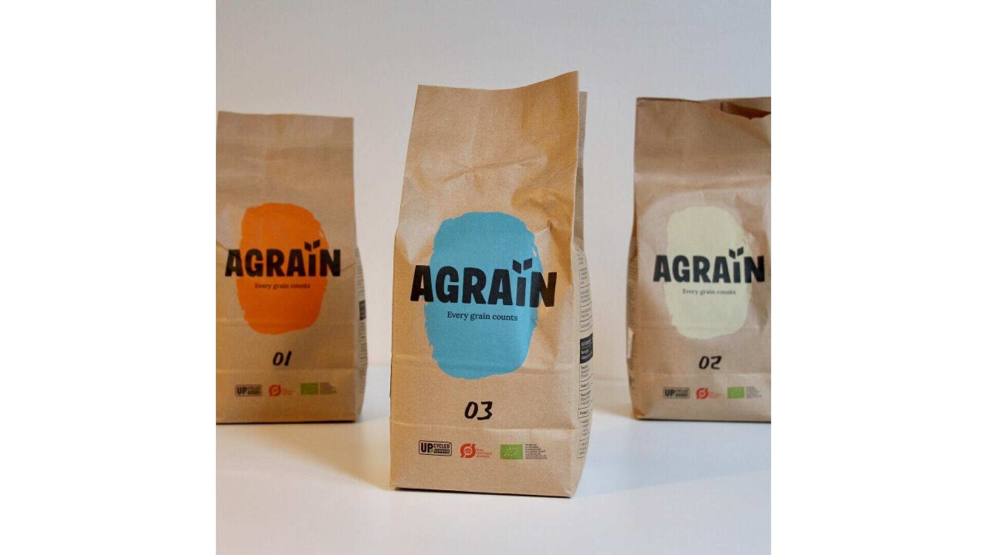 Drei braune Papiertüten mit Agrain-Mehl, jede mit einem farbigen Etikett (Orange, Blau und Creme) und den Nummern 01, 03 und 02, stehen auf einer weißen Fläche vor einem schlichten Hintergrund.