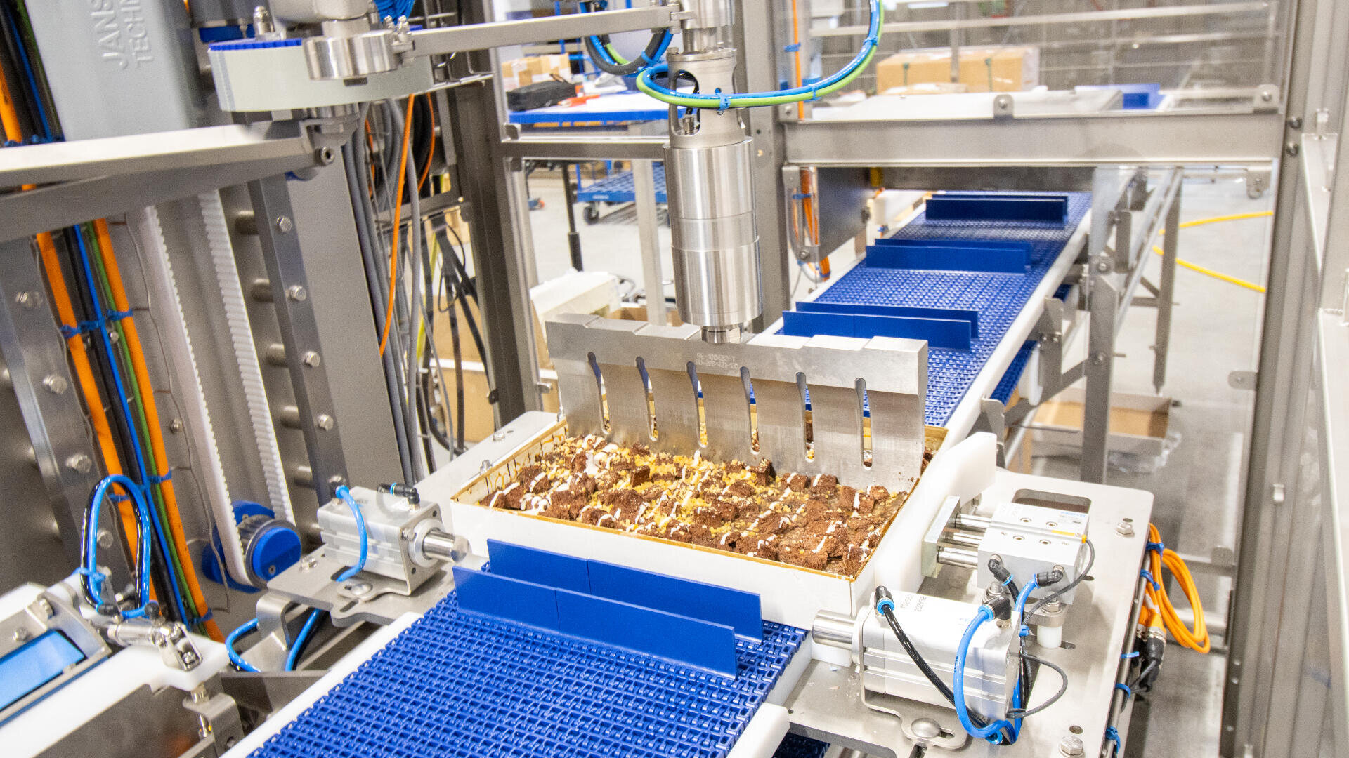 Automated factory machine with blue conveyor belts processes and slices rows of chocolate bars or snacks, using multiple blades in a bright, industrial setting.