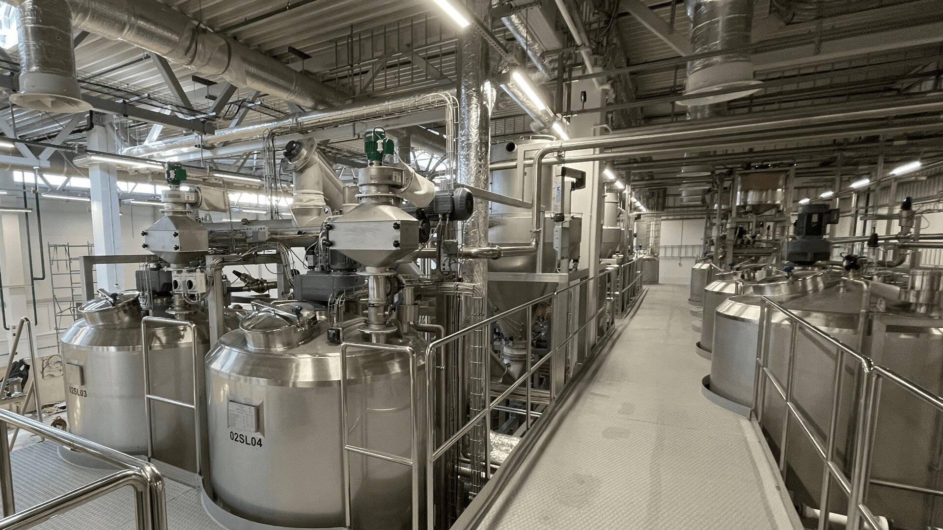 A modern industrial facility with large stainless steel tanks, pipes, and valves connected by walkways and railings, used for processing or manufacturing liquids. The setting is well-lit with overhead fluorescent lights.