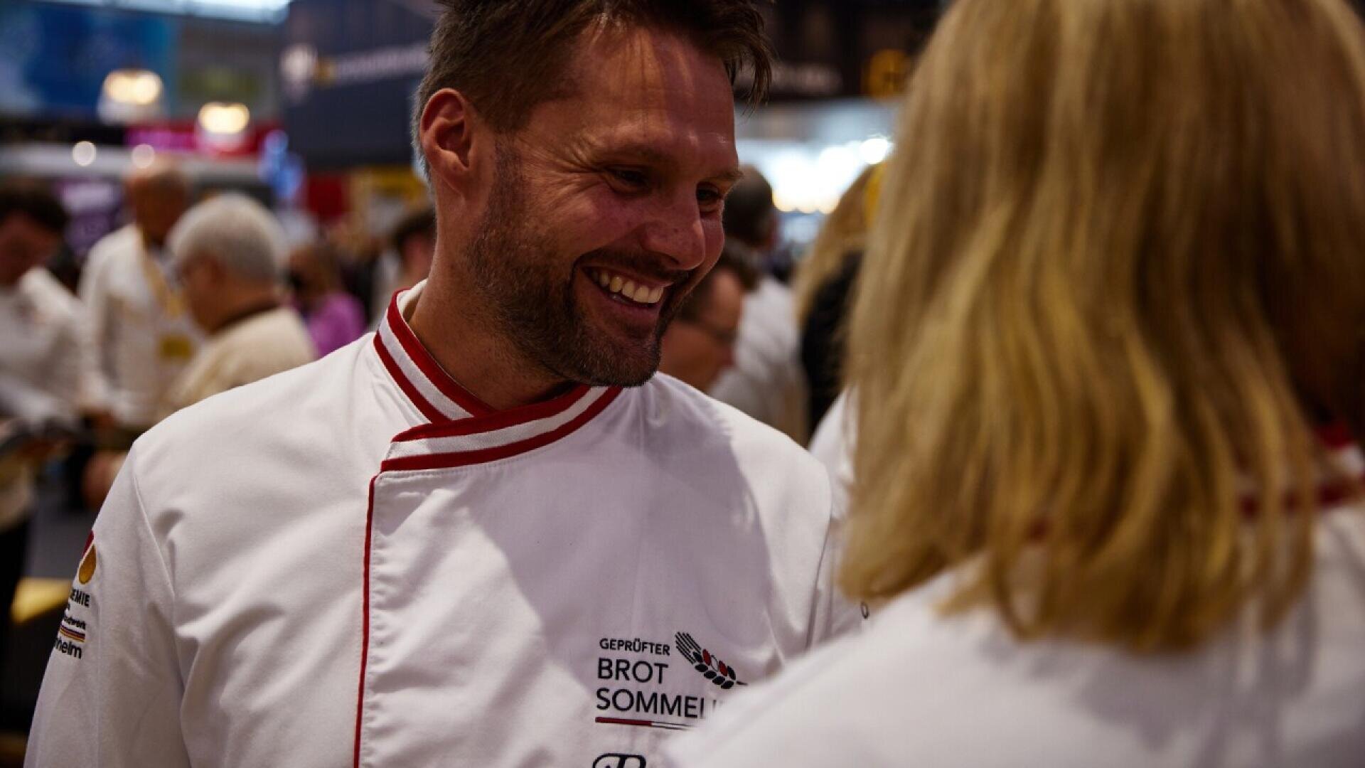 A smiling man wearing a white chefs jacket with red trim talks to a person with blonde hair in a crowded indoor setting. The jacket reads Brot Sommelier.