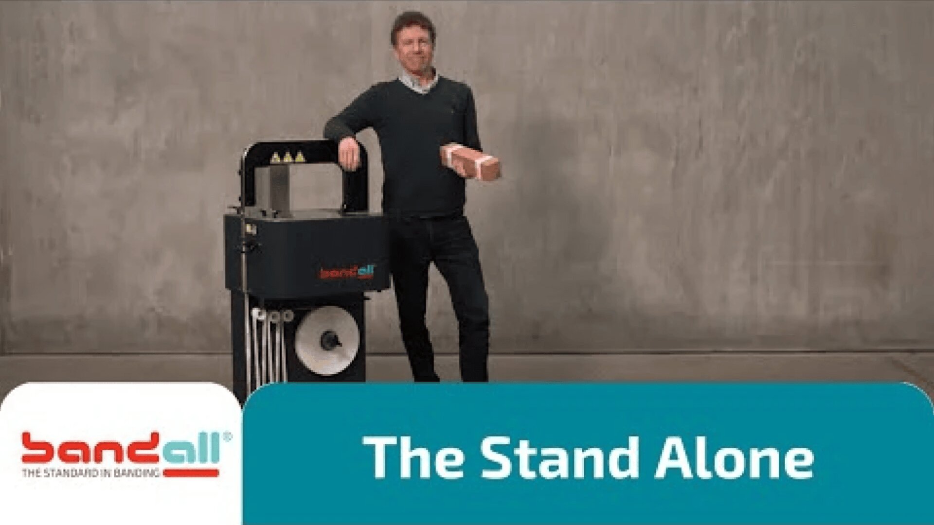 A man stands next to a Bandall strapping machine, holding a brick-shaped object. The background is plain gray, and text on the image reads, “The Stand Alone.” The Bandall logo appears in the lower left corner.