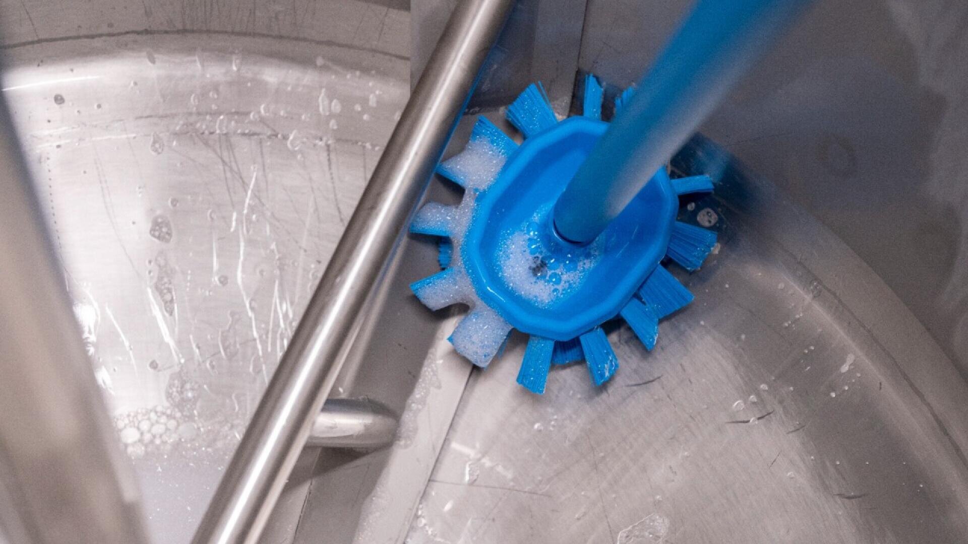 A close-up of a blue brush scrubbing the inside of a shiny metal container, with some water and soap suds visible on the surface.