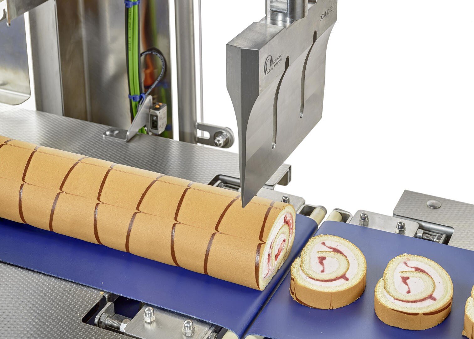 Eine Maschine mit einem großen Messer schneidet einen gerollten Kuchen mit roter und weißer Füllung auf einem blauen Förderband in gleichmäßige Stücke.