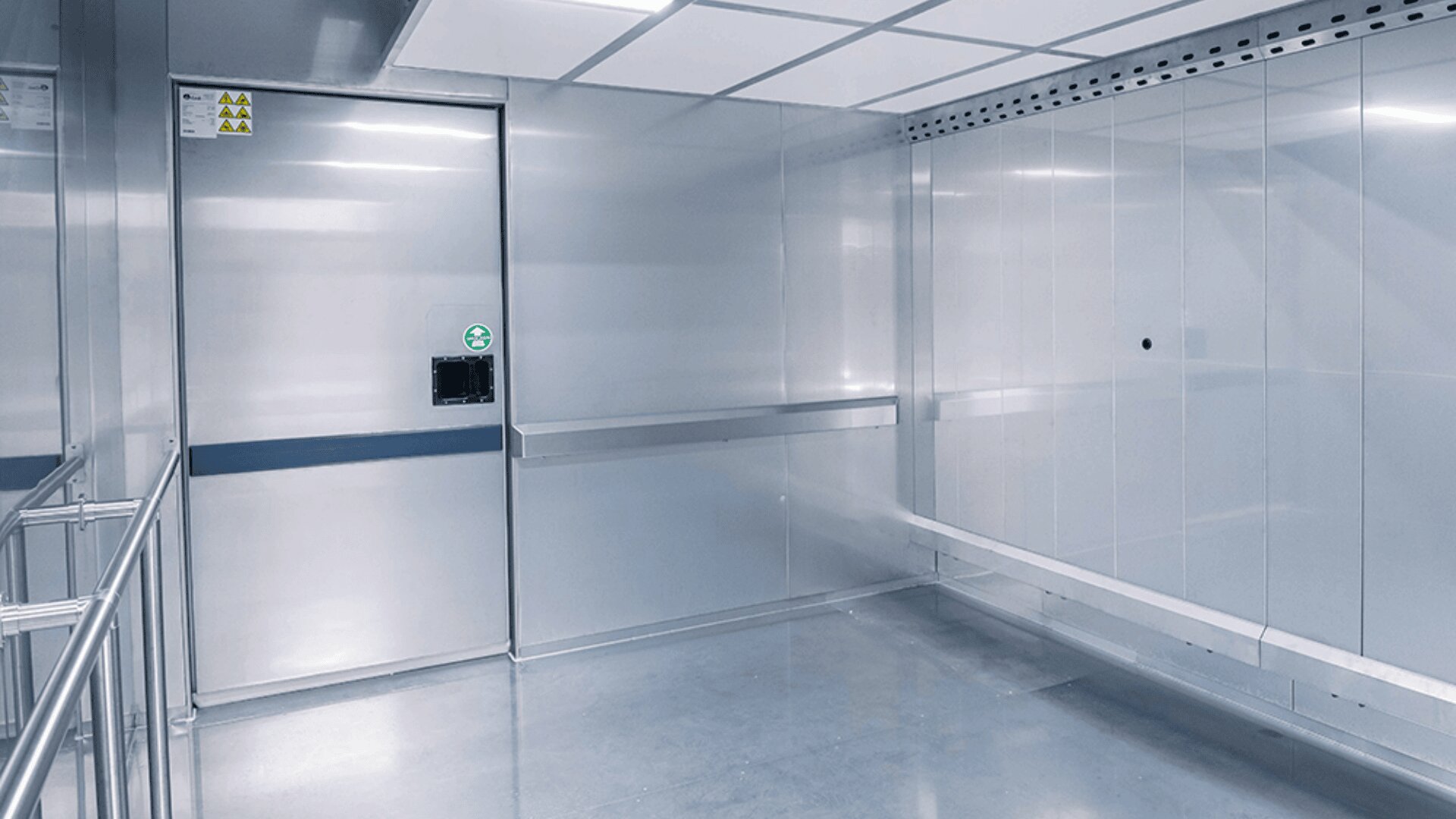 A clean, empty, stainless steel room with metal walls, a sliding door, ceiling lights, and safety signs, resembling an industrial or laboratory environment.