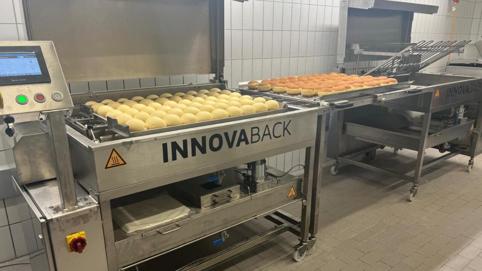 Rows of dough balls and baked bread rolls on metal trays inside an industrial bread baking machine labeled INNOVABACK in a commercial kitchen with tiled walls.