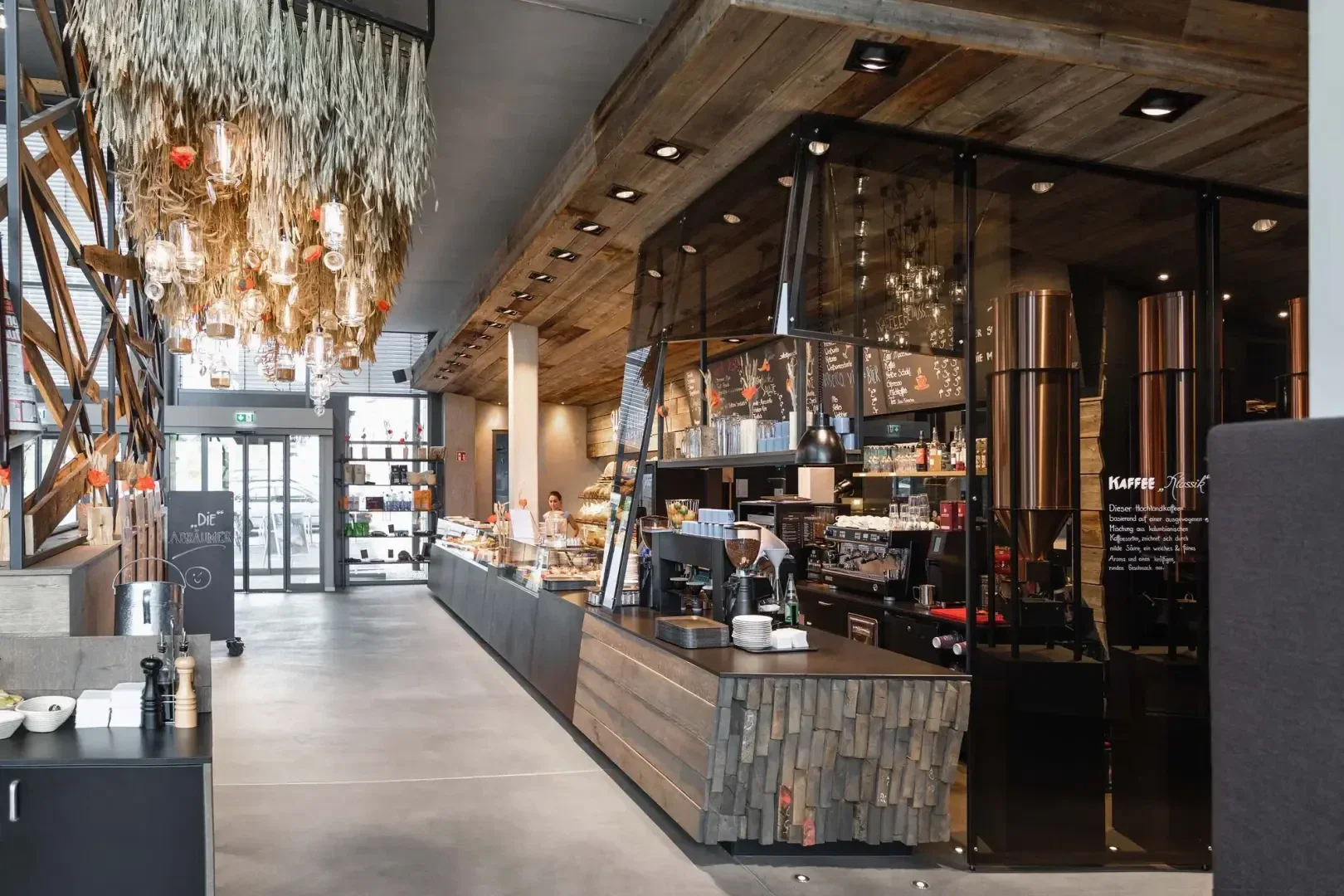 Modern café interior with a long counter, coffee machines, pastries on display, and rustic wooden accents. Hanging dried plants decorate the ceiling. Staff are behind the counter and large windows let in natural light.