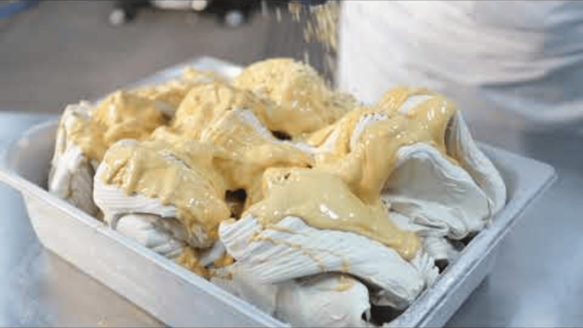 Ein mit cremigem, weißem Eis gefüllter Metallbehälter wird mit einer dicken, goldenen Soße überzogen, die von oben mit zerstoßenen Nüssen bestreut wird.