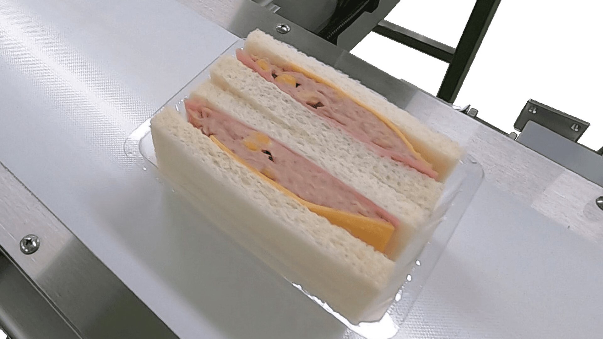 Zwei dreieckig geschnittene Sandwiches mit Weißbrot, Schinken, Käse und eventuell Ei, die in einem durchsichtigen Plastikbehälter auf einem Förderband platziert sind.