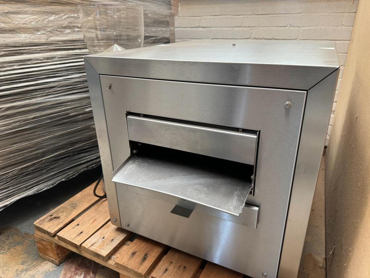 Eine Industriemaschine aus Edelstahl mit einem horizontalen Schlitz und einem Metalltablett steht auf einer Holzpalette in einem Lagerhaus, neben einem Stapel plastikverpackter Waren und einer weißen Ziegelwand.