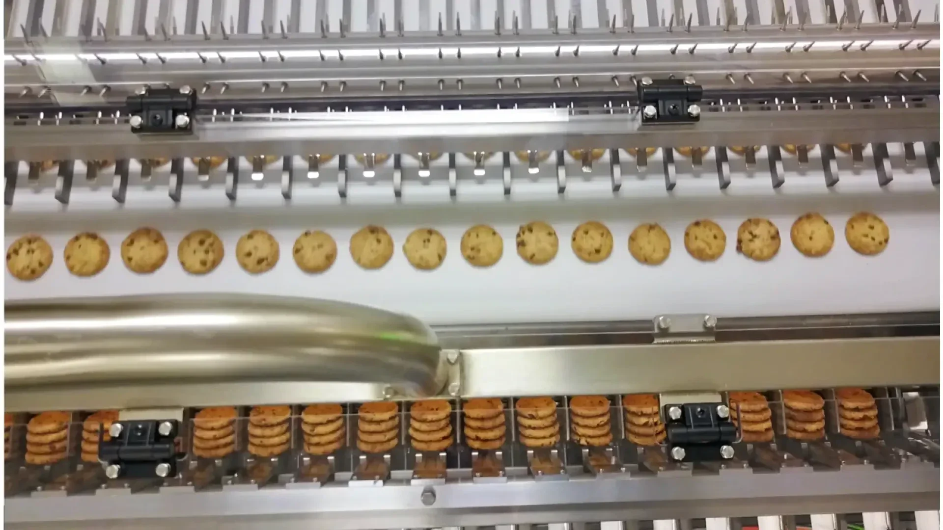 Rows of chocolate chip cookies move along a conveyor belt in a factory, passing through a section of machinery as part of an automated production process.