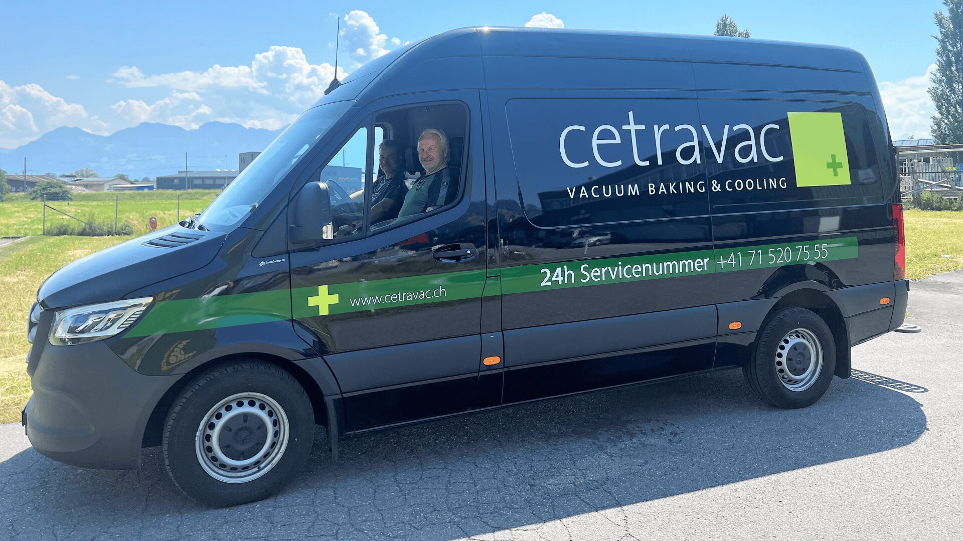 Ein schwarzer Cetravac-Kleintransporter mit grünem Schriftzug parkt an einer sonnigen Straße. Zwei Personen sitzen auf den Vordersitzen, lächeln und zeigen die Daumen nach oben. Im Hintergrund sind Berge und grasbewachsene Felder zu sehen.