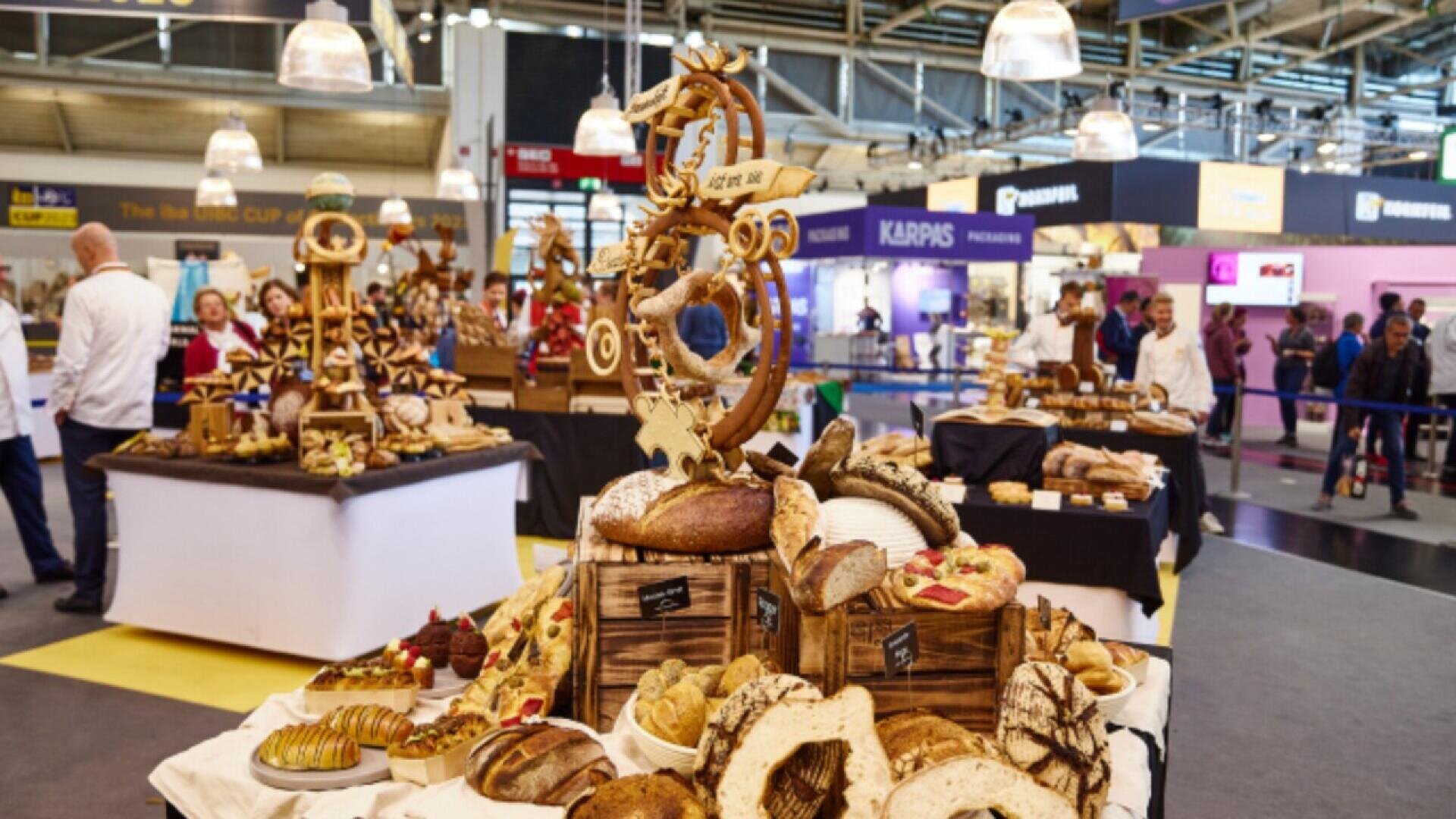 A bakery competition display shows assorted breads, pastries, and decorative bread sculptures on tables in a large indoor venue, with people walking and observing in the background.