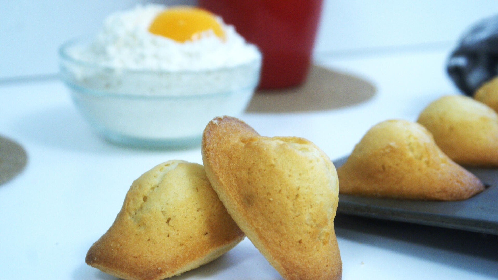 Two golden, shell-shaped madeleine cookies rest on a white surface near a baking tray; in the background, a glass bowl holds whipped cream topped with an egg yolk.