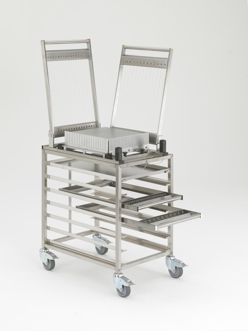 Eine Drahtschneidemaschine aus rostfreiem Stahl mit verstellbaren Rahmen ist auf einem fahrbaren Gestell montiert. Das Gerät hat freiliegende Drähte und verschiebbare Schalen, die zum Schneiden von Materialblöcken in gleichmäßige Scheiben dienen.