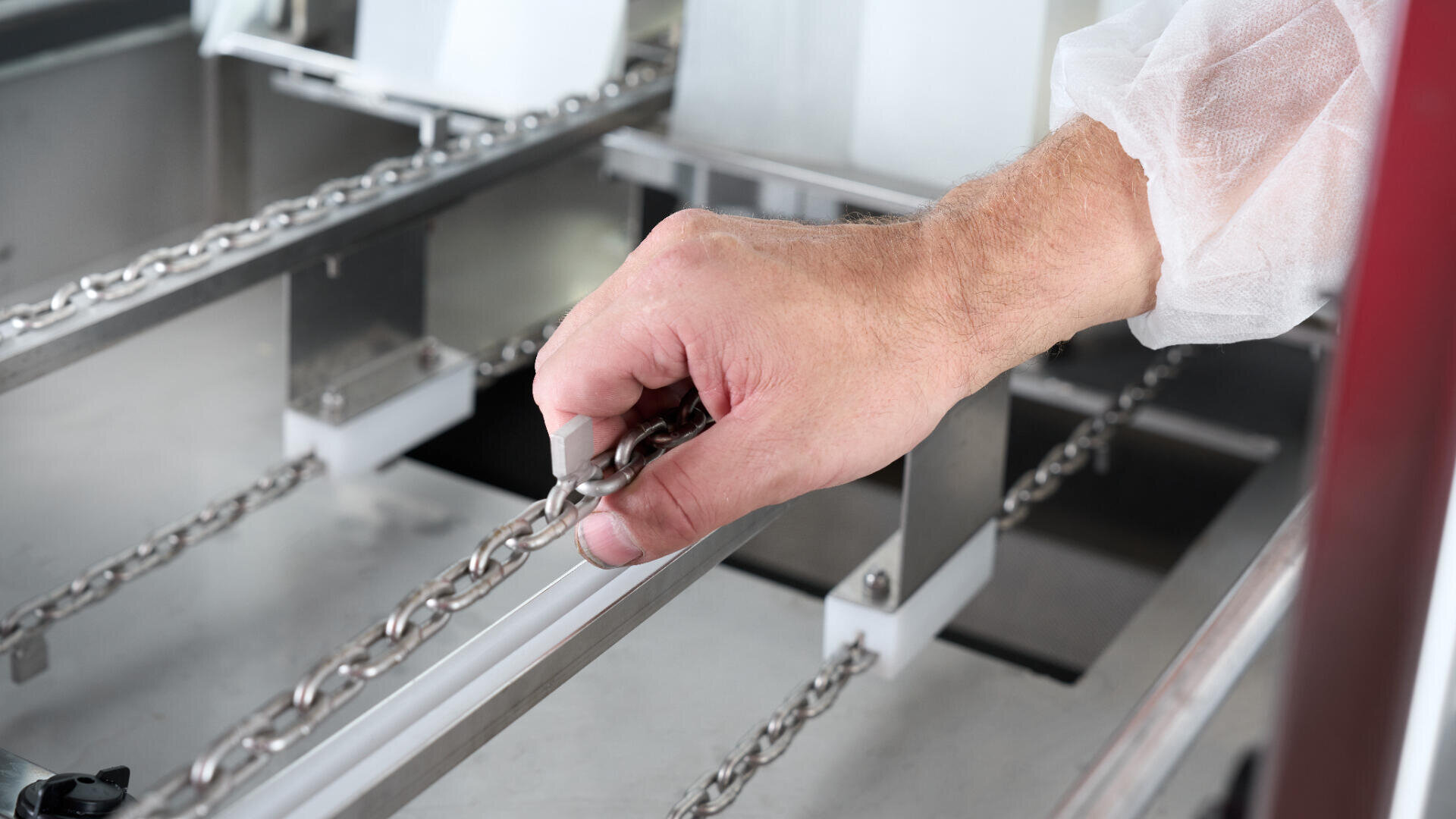 Eine Person, die einen weißen Schutzmantel trägt, prüft oder justiert eine Metallkette an einer Industrieanlage oder Maschine.