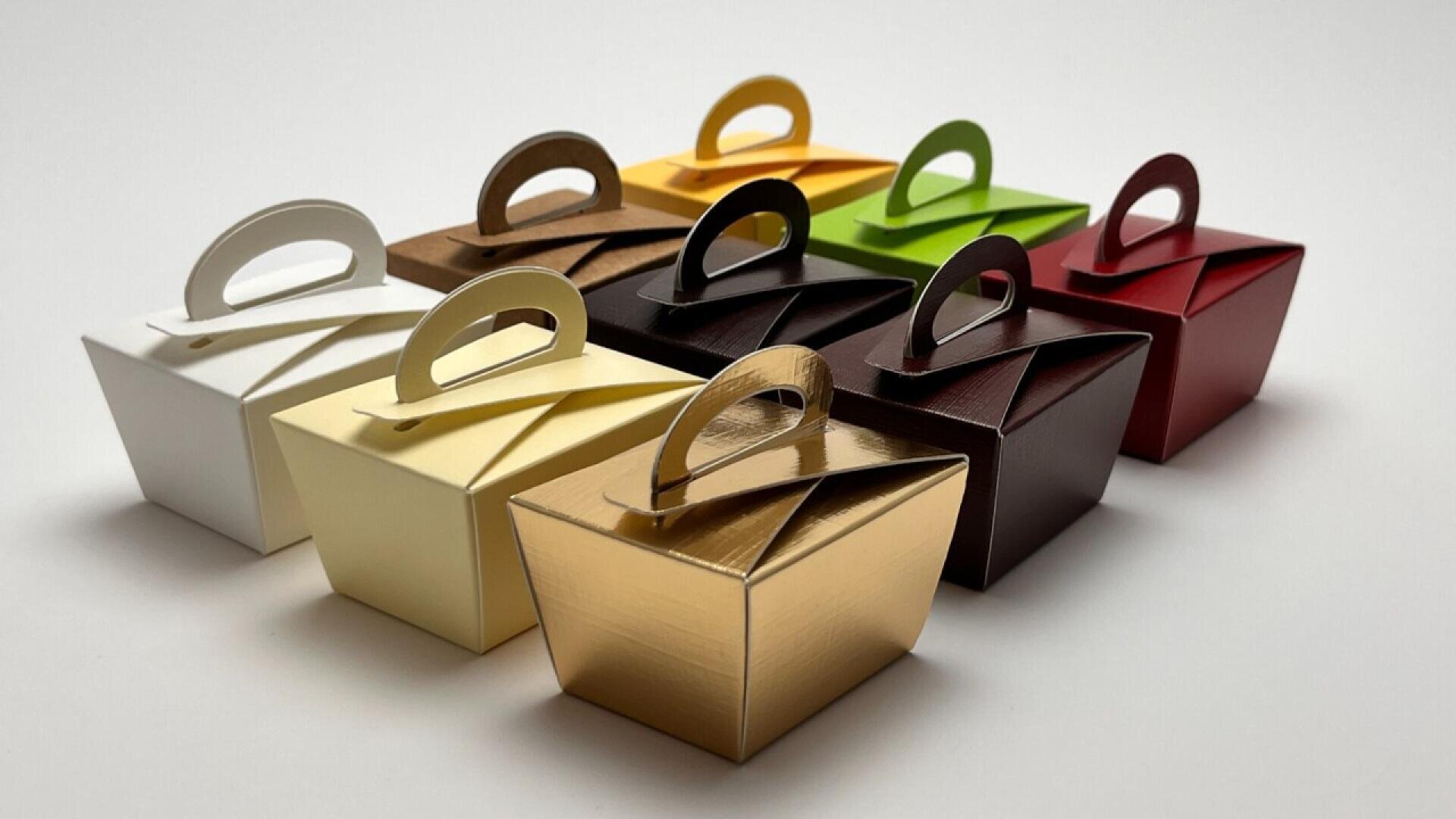 Eight small, glossy gift boxes with handles are neatly arranged in two rows. Each box is a different color, including white, yellow, gold, bronze, black, maroon, green, and red, on a plain white background.