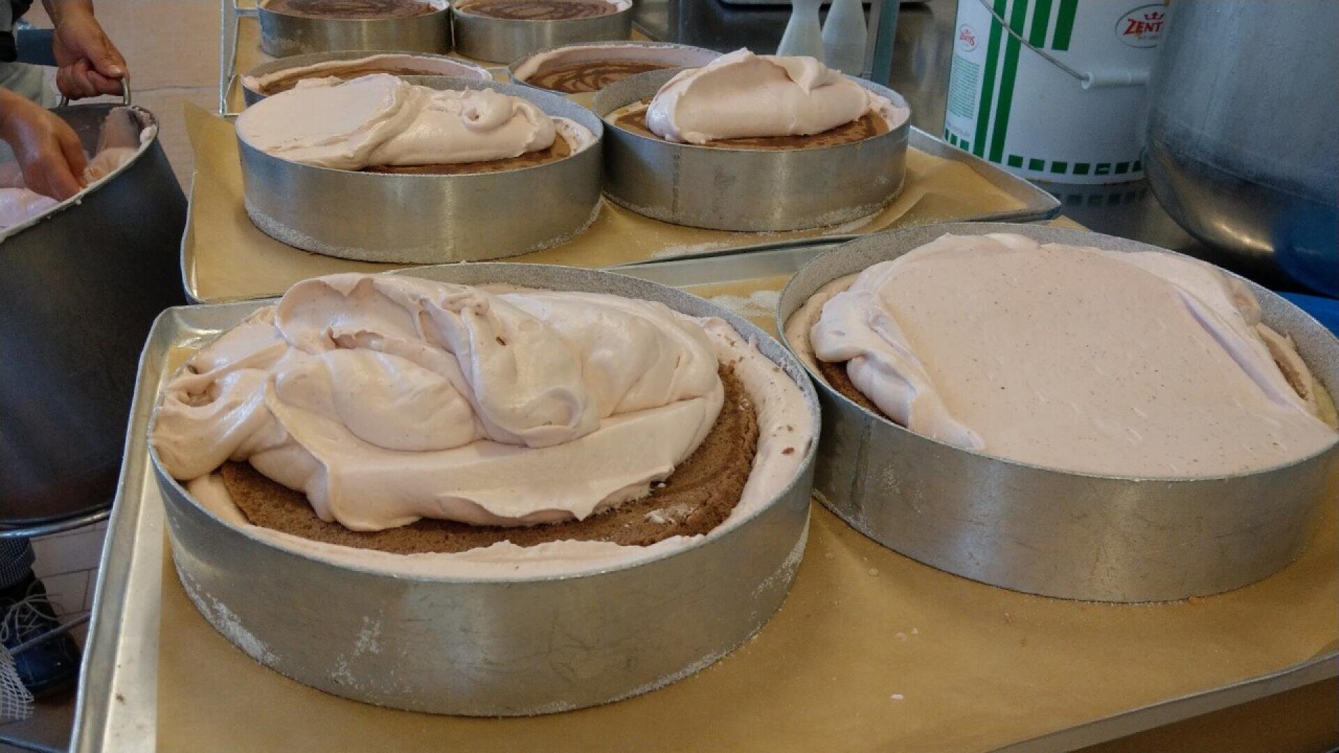 Round baking pans filled with cake layers topped with thick, light pink whipped frosting, placed on a tray in a kitchen setting. A person’s hand is visible on the left, working with the cakes.