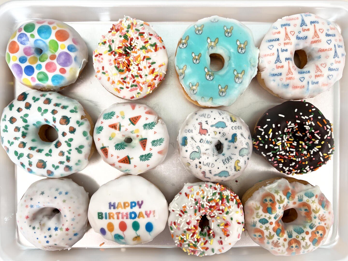 Ein Tablett mit zwölf dekorierten Donuts, jeder mit farbenfrohem Zuckerguss und einzigartigen Motiven wie Streuseln, Tupfen, Tieren, Happy Birthday und verschiedenen verspielten Mustern.
