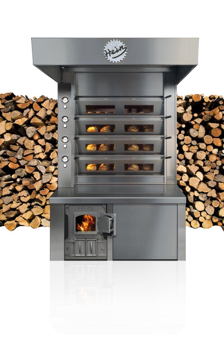Ein moderner holzbefeuerter Ofen aus Edelstahl mit fünf Backkammern, die mit Brotlaiben gefüllt sind, steht vor ordentlich gestapelten Holzscheiten.