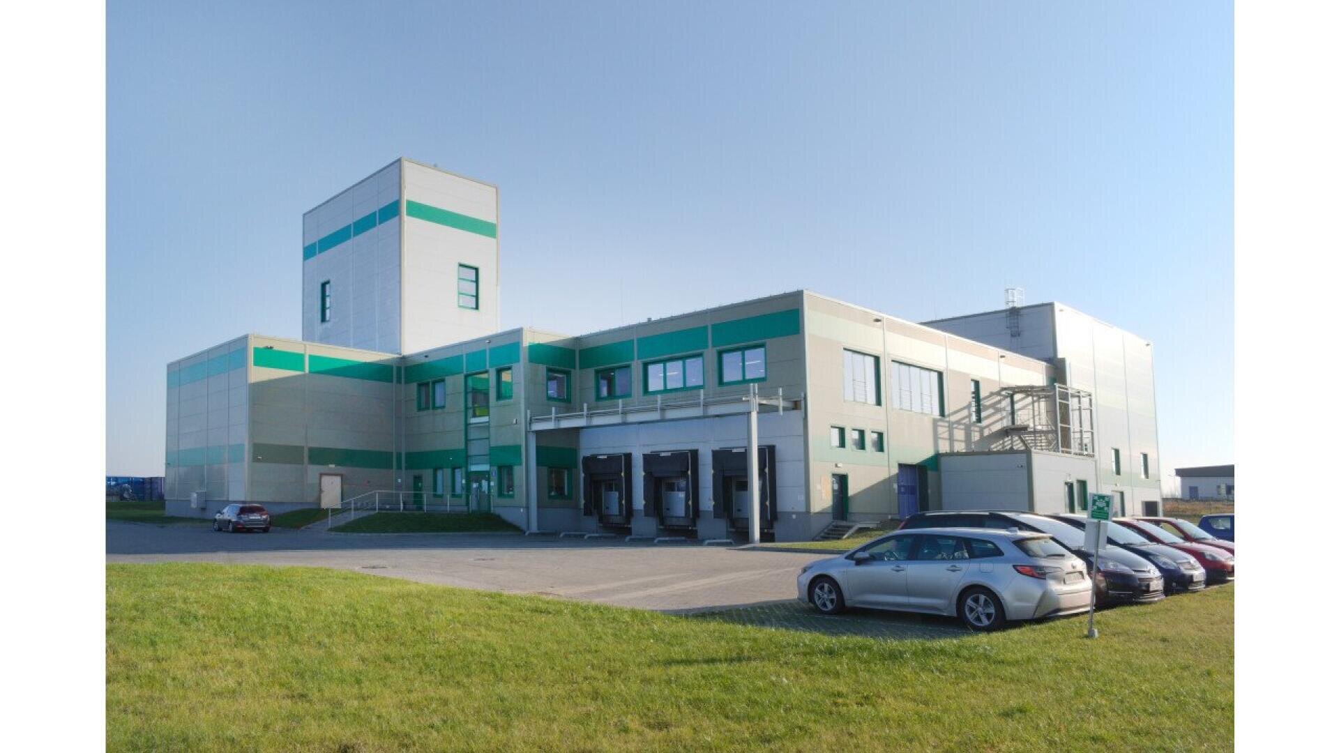 Ein modernes Industriegebäude mit grünen und grauen Wänden, Ladedocks und mehreren davor geparkten Autos an einem sonnigen Tag, mit einer Grünfläche im Vordergrund.
