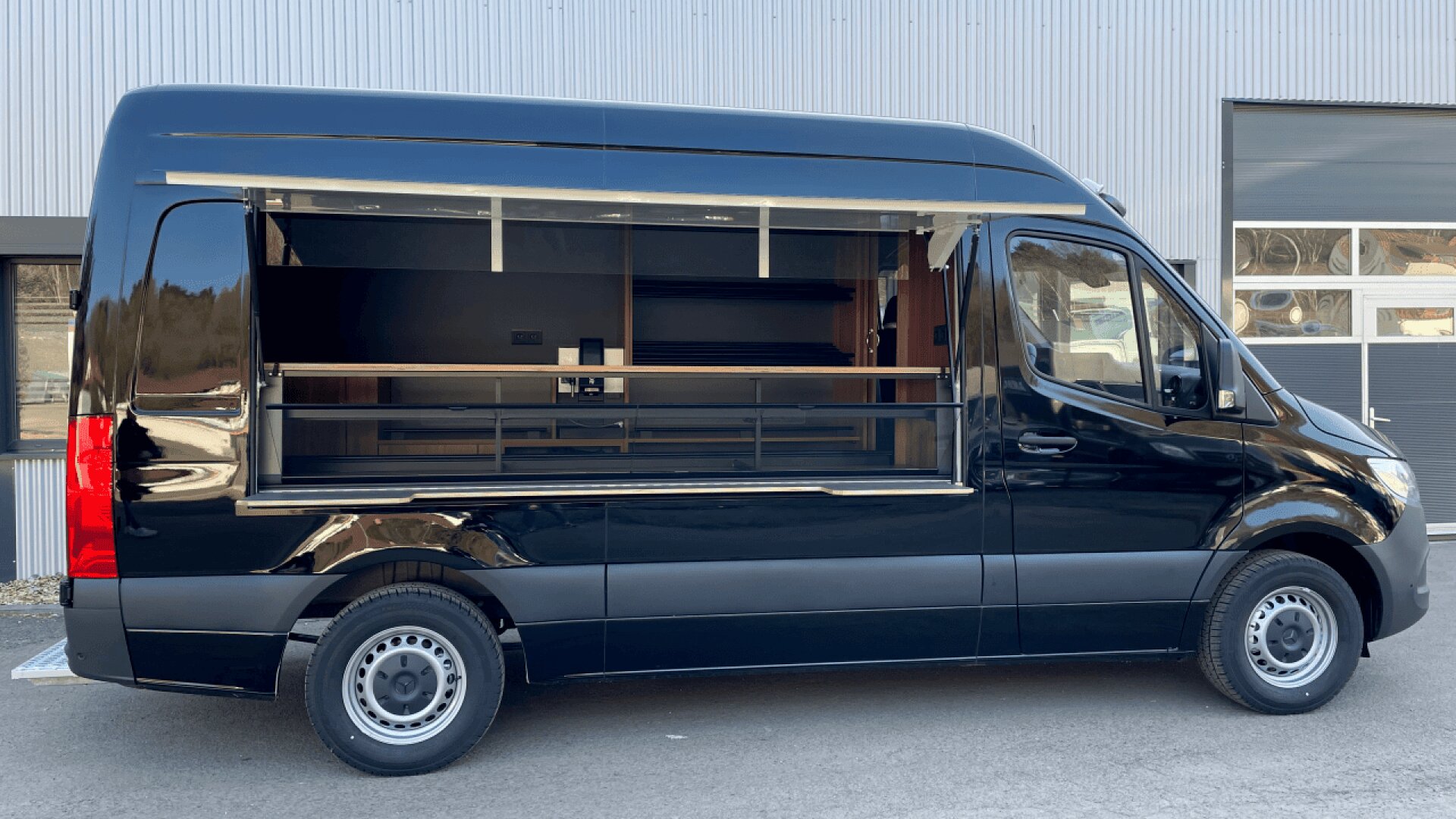 A black food truck with an open service window is parked outside a building with gray metal siding and large windows.