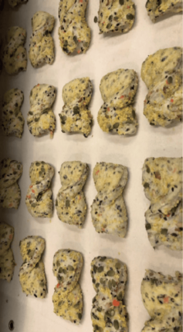 Auf einem mit Pergamentpapier ausgelegten Backblech liegen Reihen von rohen, rechteckigen, mit Samen und Kräutern gefüllten Teiglingen, die zum Backen bereit sind.